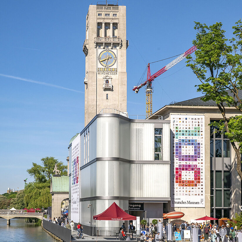Blick auf den Uferbereich des Deutschen Museums in München: im Vordergrund moderne Glasfassade und Besucherpavillon am Fluss mit Menschen auf der Promenade, rechts bunte Plakate an der Museumsfassade, im Hintergrund der hohe Turm mit großem Zifferblatt und der Aufschrift „WINDMESSER“, ein roter Baukran und blauer Himmel.