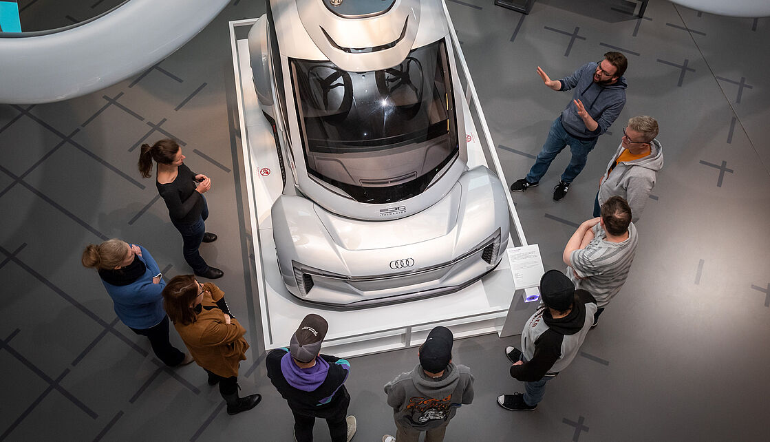 Ein silbernes Audi-Studienfahrzeug auf weißem Podest in einer Ausstellung, umgeben von Besuchern aus der Vogelperspektive — moderne Karosserie mit großem Panorama-Glasdach, dezenter Marken-Logos vorne, Infotafel rechts; glatter grauer Ausstellungsboden mit Kreuzmustern.