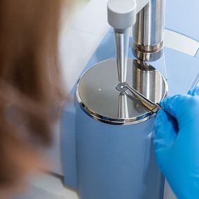 Nahaufnahme eines Laborgeräts: eine Person mit blauen Einmalhandschuhen hält feine Pinzetten und positioniert eine kleine Probe auf einer glänzenden Metallplatte unter einer präzisen Einstich- oder Messsonde an einem hellblauen Prüfgerät.