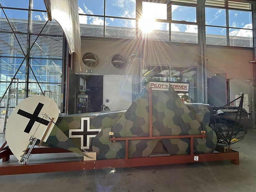 Rumpfabschnitt eines historischen Flugzeugs mit sechseckiger Tarnlackierung in Grün-, Braun- und Olivtönen auf einem roten Transportgestell, sichtbares Heck mit schwarzem Balkenkreuz auf weißem Kreis, im Hintergrund große Hallenfenster mit Sonnenlicht und Schild „Pilot’s Corner“.