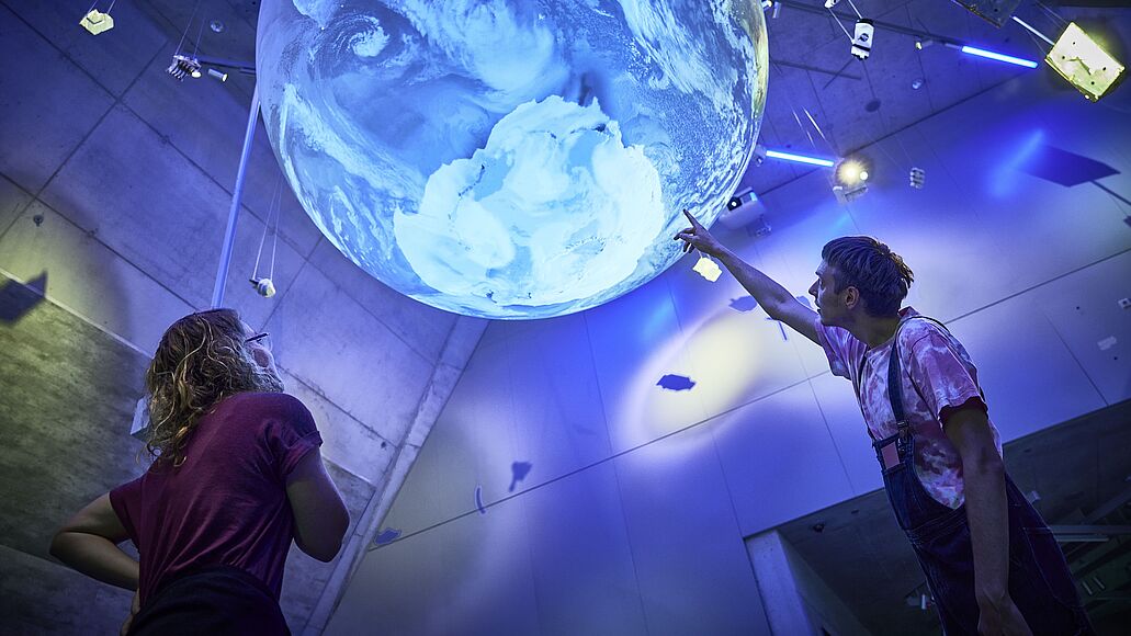 Zwei Besucher in einem dunklen Museumssaal betrachten und zeigen auf eine große, leuchtende Erdkugel-Installation, die von der Decke hängt; blaues Licht, Betonwände, eine Person links mit roten T‑Shirt und lange Haare, rechts eine Person in Latzhose, die mit ausgestrecktem Arm auf die Darstellung der Südpol-Region zeigt.