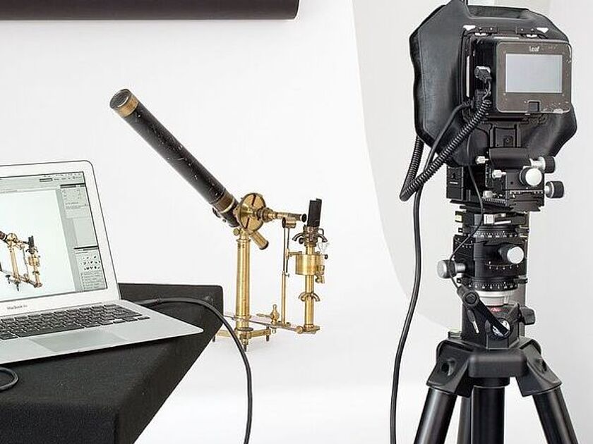 Studioaufnahme eines antiken Messinstruments (Brass-Teleskop oder wissenschaftliches Messgerät) auf einem Stativ, fotografiert mit einer professionellen Großformatkamera auf Dreibein; links ein Laptop zeigt das Live-Bild des Objekts, Hintergrund weißer Fotohintergrund und oberhalb gerolltes schwarzes Papier.