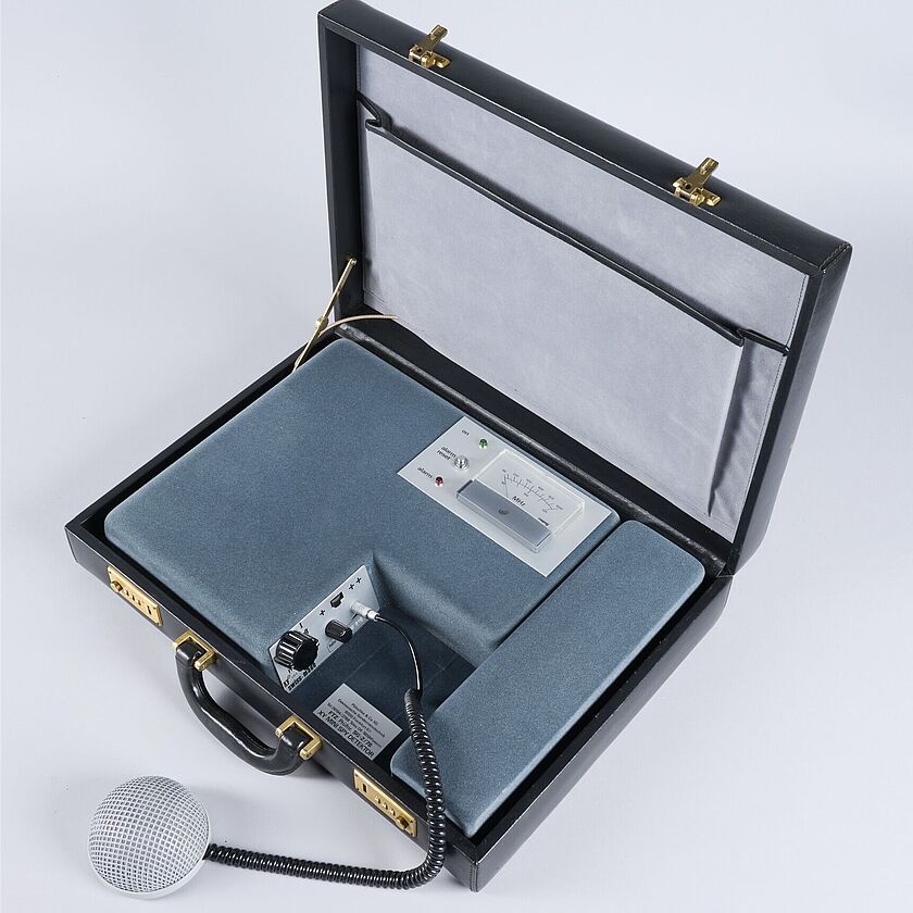 2025-135_20250617_002_SAMOA_ND_AKaufmann.jpg Schwarze, geöffneter Lederkoffer. Darin eingelegt: mit Samt umzogener Apparat mit MHz Anzeige, drei Lämpchen und einem Mikrofonanschluss mit Reglern.