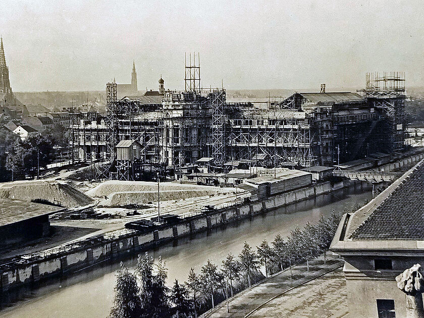 Schwarzweiß-Foto einer großen Baustelle an einem Flussufer: imposantes historisches Gebäude im Gerüstaufbau mit Türmen, Gerüsten und Bauhütten, davor aufgeschüttete Sandflächen, Kaimauer und Wasserkanal; im Vordergrund ein schräges Dach mit Ziegeln und ein steinernes Dekorationsstück, im Hintergrund Stadtsilhouette mit zwei markanten Kirchtürmen unter hellem Himmel.