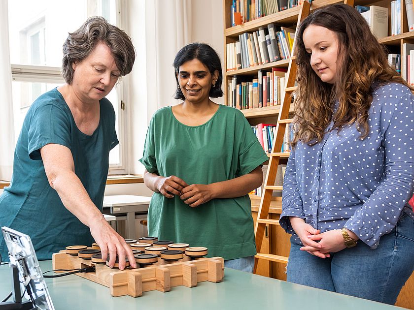 Drei Frauen stehen an einem Tisch in einer Bibliothek; links eine Frau greift auf ein hölzernes Spiel- oder Experimentierbrett mit mehreren runden Scheiben, in der Mitte eine Frau in grünem Shirt, rechts eine Frau in blauem, gepunktetem Hemd; im Hintergrund Bücherregale und eine Leiter.