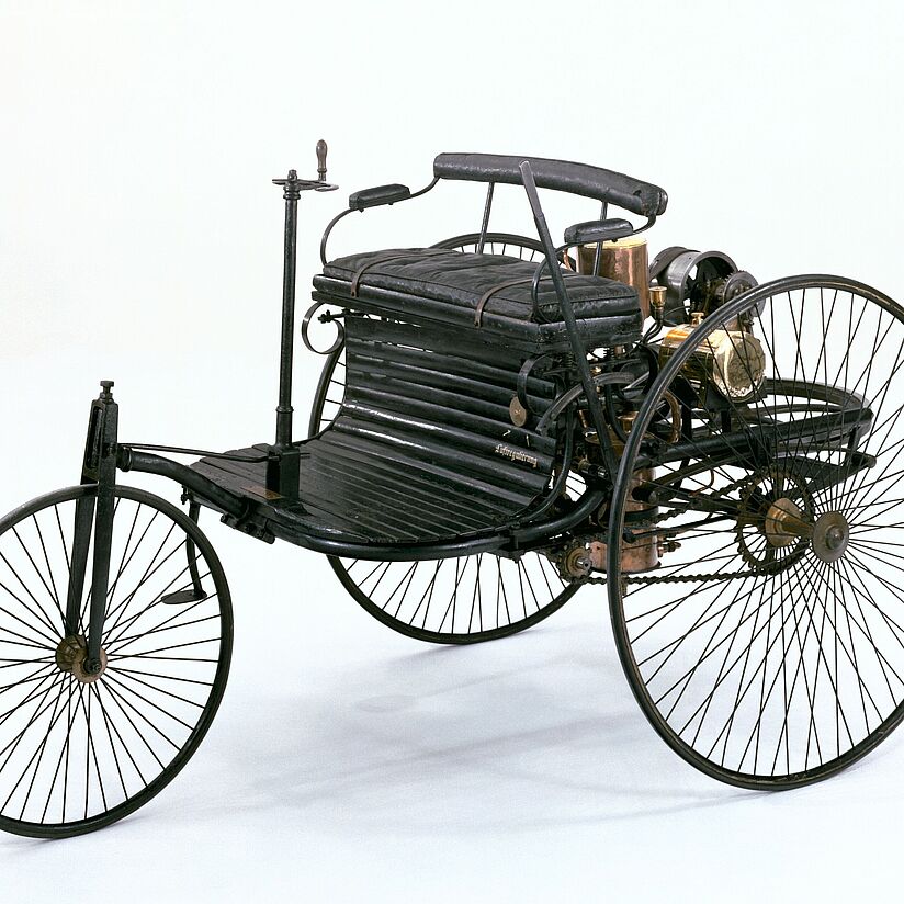 Ein frühes dreirädriges Automobil (Benz Patent-Motorwagen) mit schmalen Metall-Speichenrädern, gepolstertem Zweiersitz, sichtbarem Messingmotor und Kettenantrieb vor weißem Hintergrund.