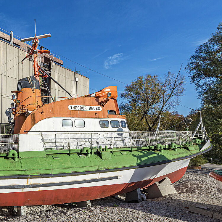 Rettungsboot „Theodor Heuss“ in Orange, Weiß und Grün auf Landgestell vor einem Betongebäude bei klarem Himmel, seitliche Ansicht mit Decksgeländer und Rettungsinseln.
