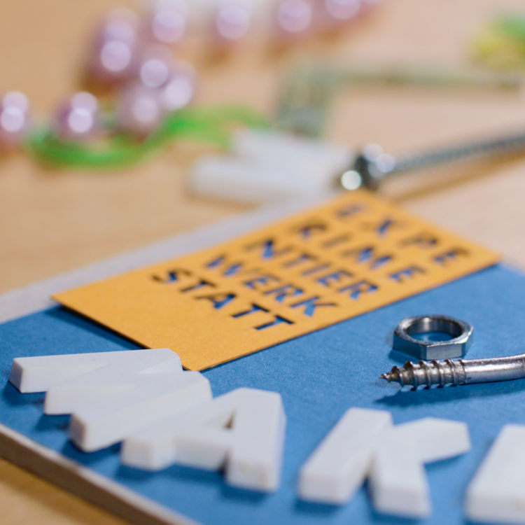 Nahaufnahme einer kreativen Werkstatt-Szene. Im Vordergrund sind eine Schrauböse, ein Metallring und weiße Buchstaben zu sehen, die das Wort „MAKE“ bilden. Im Hintergrund liegt ein orangefarbenes Papierstück mit der Aufschrift „EXPERIMENTIERWERKSTATT“. Verschiedene kleine Bastelmaterialien wie Perlen und Schrauben sind ebenfalls sichtbar.