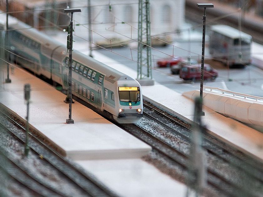 Detail der Modelleisenbahn. Zu sehen sind der Bahnhof mit Parkplatz, zwei überdachte Bahnsteige und ein einfahrender Zug.