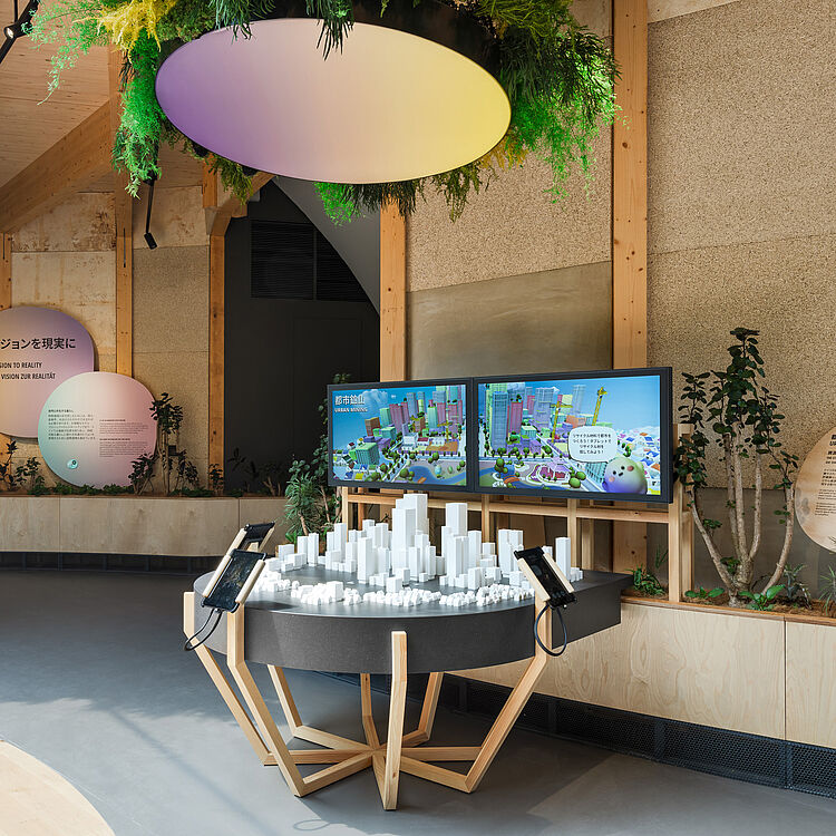 Interaktives Ausstellungsdisplay mit einem runden Tisch auf hölzernem Gestell, darauf ein weißes 3D-Modell einer Stadt; zwei Bildschirme im Hintergrund zeigen farbige Stadtgrafiken; große ovale Deckenleuchte mit hängender Begrünung, Pflanzen und erklärende Texttafeln an den Wänden.