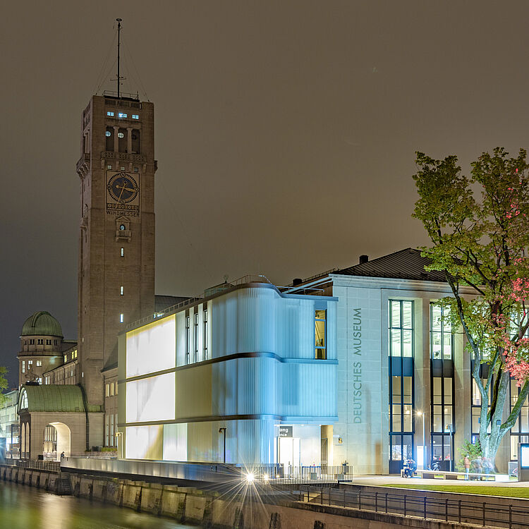 Das nächtlich beleuchtete Deutsches Museum am Flussufer mit modernem, blau beleuchteten Neubau, hohem Uhrenturm mit Aufschrift „Windmesser“, großen Bäumen rechts und Reflexionen im Wasser.