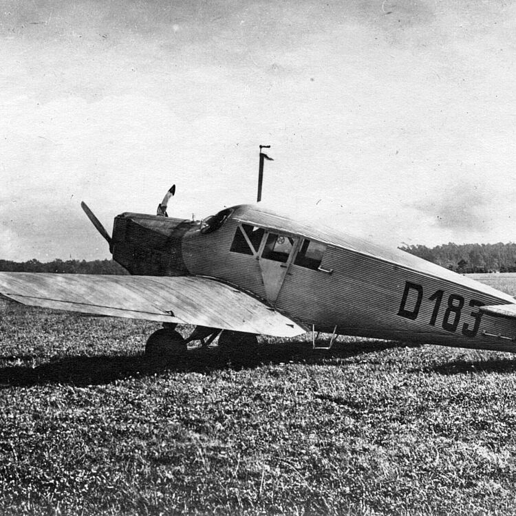 Junker F 13 Schwarzweiß-Foto eines einmotorigen Landflugzeugs mit Kennung "D183" auf dem Rumpf, geparkt auf einer Grasfläche; Seitenansicht zeigt einen starren Tiefdecker mit zweiblättriger Holzpropellernase, geschlossener Kabine mit rechteckigen Fenstern, spitz zulaufendem Heckleitwerk und sichtbaren Reifen im Fahrwerk vor einer baumbestandenen Horizontlinie.