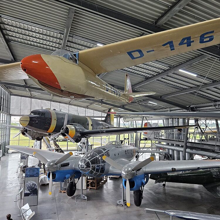 Innenansicht eines Luftfahrtmuseums mit mehreren ausgestellten Flugzeugen: vorne ein zweimotoriges, silber-blau lackiertes Propellerflugzeug, darüber ein dunkles Transportflugzeug mit gelb-schwarzen Triebwerksverkleidungen und im Vordergrund hängend ein heller Segler mit roter Nase und der Kennung D-1469 an der Tragfläche; große Hallenfenster, Stahlträger und Besucher am Boden sind sichtbar.