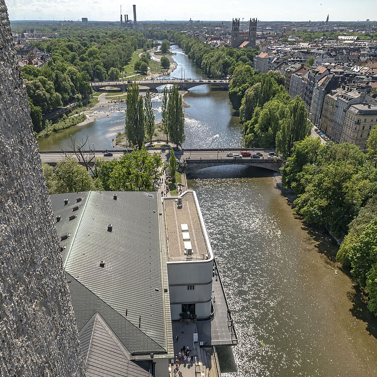 Blick aus der Vogelperspektive auf einen Fluss mit glitzernder Wasseroberfläche, rechts von Bäumen gesäumtes Flussufer, links ein großes Gebäudedach und eine Promenade mit Spaziergängern; mehrere Brücken überspannen den Fluss, im Hintergrund Parklandschaft und Stadt mit Kirchen- und Fabrikgebäuden am Horizont.