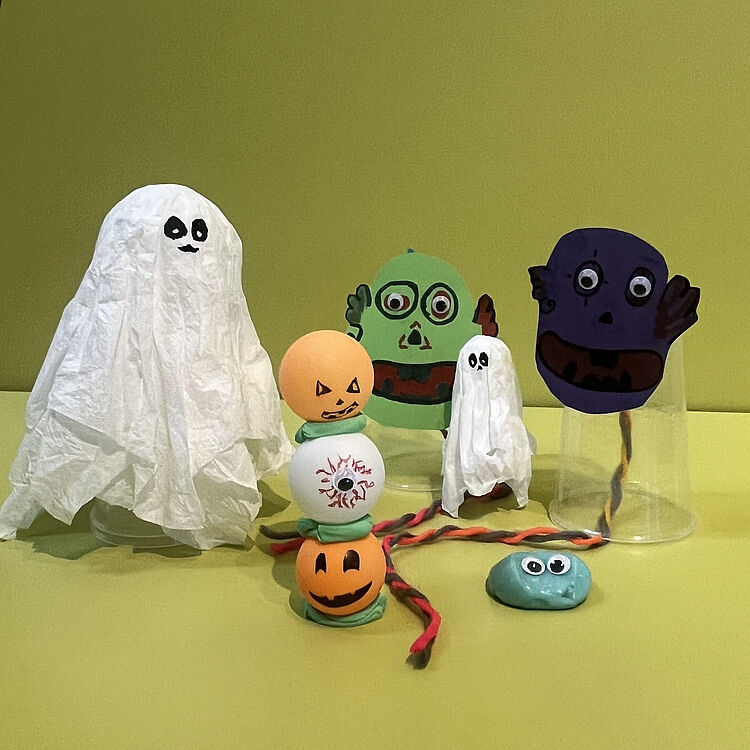 Mehrere gebastelte Halloween-Figuren: links ein großes Gespenst, vorne ein Kürbisturm-Turm aus drei bemalten Tischtennisbällen, daneben kleines Taschentuchgespenst, hinten zwei grüne und violette Monster mit aufgeklebten Wackelaugen.