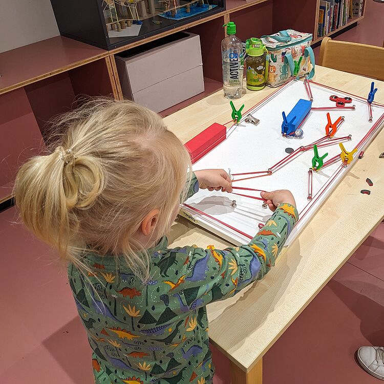 Kind spielt an einem selbstgebauten Flipper-Tisch aus Alltagsmaterialien mit bunten Klammern, Gummibändern und Magneten.