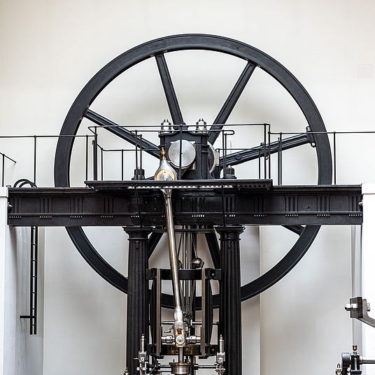Großes schwarzes Schwungrad und historische Dampfmaschine mit sichtbarem Pleuel, Kolben und Gusseisenrahmen in einem hellen Museumsraum, neben einer Ziegelmauer und Balustrade.