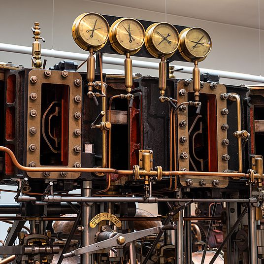 Nahaufnahme einer historischen Dampfmaschine mit sichtbaren Zylindern, Messingarmaturen und kupfernen Rohrleitungen; oben vier runde Messgeräte in Messingfassung, massive gusseiserne Gehäuse mit Schrauben, Ventilen, Hebeln und Gestänge, komplexe mechanische Verbindungen und Beschriftungspatte an einer Kurbel, Ausstellungsraum-Hintergrund.