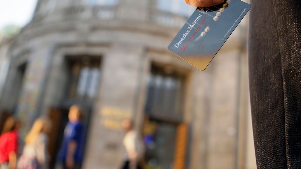 Nahaufnahme einer Hand mit Armbanduhr, die eine Mitgliedskarte des Deutschen Museums hält; unscharfes historisches Museumsgebäude und Passanten im Hintergrund.