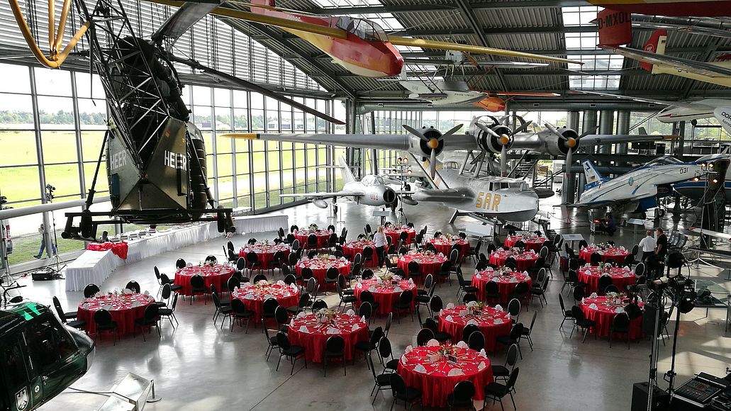 Großer Flugzeughangar, im Inneren zahlreiche historische Flugzeuge und Hubschrauber über und neben rund eingedeckten Tischen mit roten Tischdecken für eine Galaveranstaltung, Techniker und Bühnentechnik am rechten Rand.