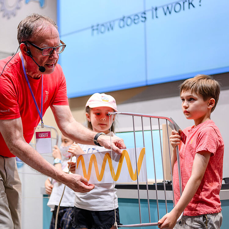 Ein Aussteller in rotem T‑Shirt hält vor zwei Kindern ein transparentes Blatt mit gelber Wellenlinie an ein Metallgitter, im Hintergrund Bildschirm mit teilweise sichtbarer Frage "How does it work?".