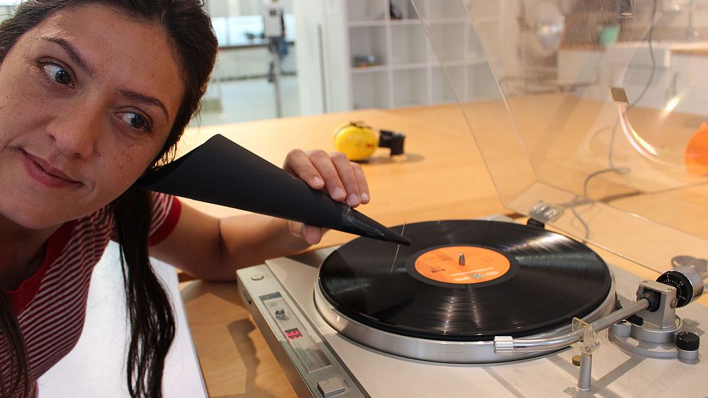 Hör-Werkstatt Frau hört mit einem Trichter aus Pappe eine Schallplatte. Der Trichter hat am Ende eine Nadel. Diese gleitet über die Schallplatte, während sie sich dreht.
