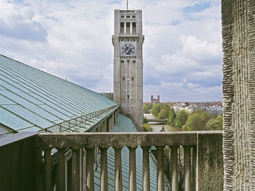 Blick von einem steinernen Balkon über grüne Kupferdächer auf einen hohen Beton-Turm mit großem Barometer-Zifferblatt und vertikaler Messskala, im Hintergrund Fluss, Bäume und Stadtgebäude unter blau-weilem Himmel.