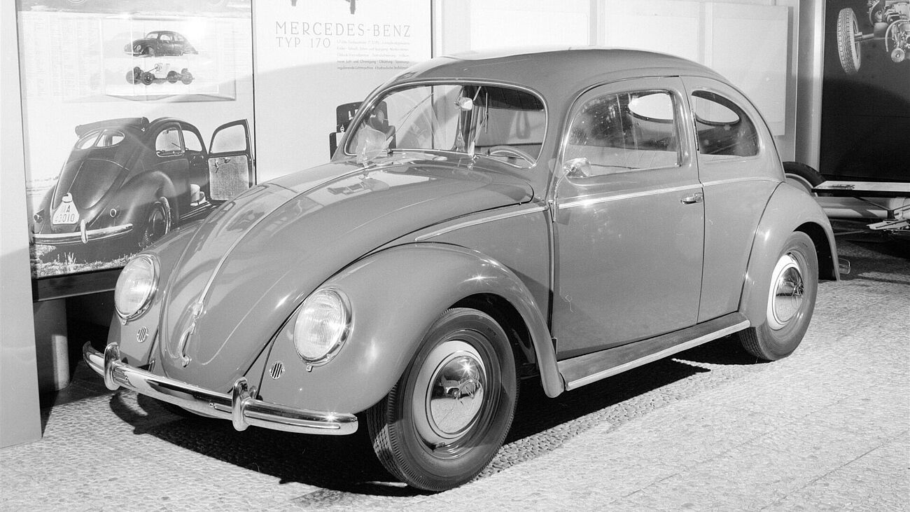 Schwarzweiß Aufnahme eines VW Käfers in den Ausstellungsräumen vor bebilderten und beschrifteten Ausstellungswänden. Das Auto ist von vorne rechts aufgenommen. Am Bildrand unten Links ist ein schwarzes Täfelchen mit der Aufschrift "L18/84", vermutlich die Inventarnummer des Käfers, zu erkennen.