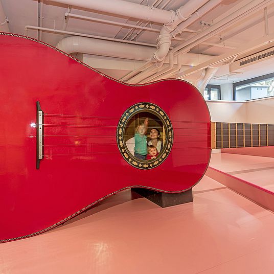 Riesengitarre Drei Kinder spielen mit der Riesengitarre. Zwei von ihnen sind in der Gitarre, ein Junge zupft von außen eine Seite.