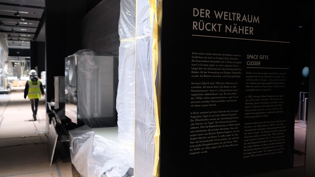 Ausstellung im Umbau. Links im Hintergrund: Person mit Warnweste und Bauhelm. Rechts im Vordergrund: Abgeklebte Wände und ein Plakat "Der Weltraum rückt näher"