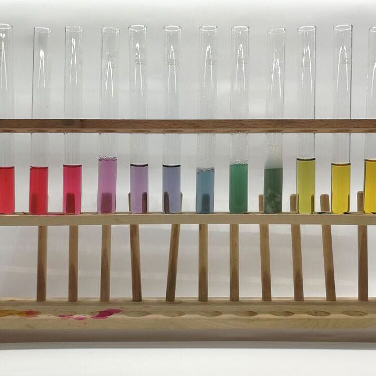 Das Bild zeigt eine Reihe von Reagenzgläsern in einem Holzständer, die Flüssigkeiten mit unterschiedlichen Farben enthalten. Diese Farben repräsentieren den pH-Wert einer Lösung, von Rot (sauer) über Lila (neutral) bis Gelb (basisch).