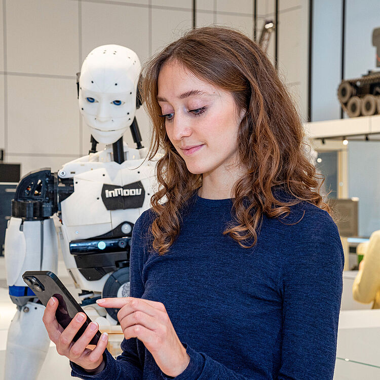 Junge Museumsbesucherin blickt auf ihr Smartfone, welches sie in der Hand hält. Im Hintergrund steht ein menschlich aussehender Roboter.