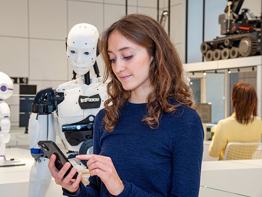 Junge Museumsbesucherin blickt auf ihr Smartfone, welches sie in der Hand hält. Im Hintergrund steht ein menschlich aussehender Roboter.