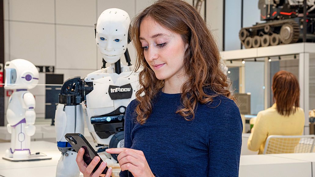Junge Museumsbesucherin blickt auf ihr Smartfone, welches sie in der Hand hält. Im Hintergrund steht ein menschlich aussehender Roboter.