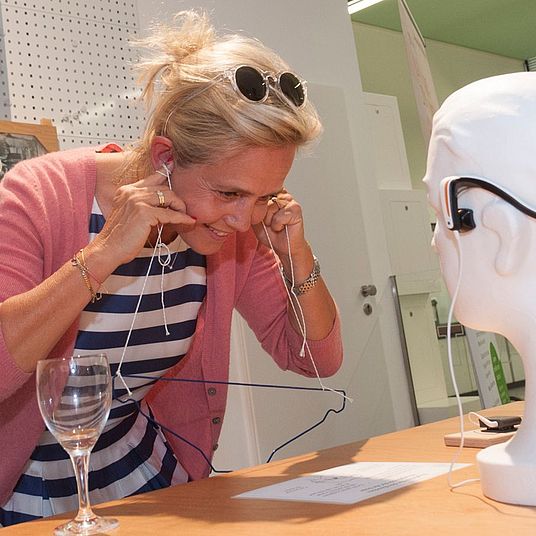 Frau mit verpixeltem Gesicht, trägt gestreiftes Kleid und rosa Cardigan, steckt Kopfhörer in die Ohren; vor ihr auf dem Tisch ein Weinglas, Drahtbügel und ein Schaumstoff-Kopf mit aufgesetzten Kopfhörern in einem hellen, gut beleuchteten Technik- oder Ausstellungsraum.