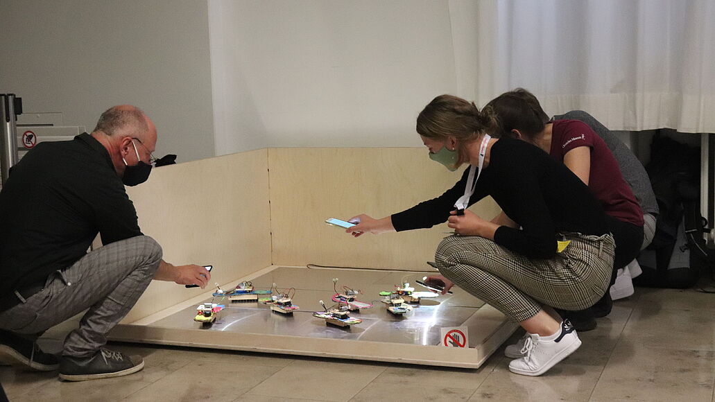 Drei Personen, hockend an einem niedrigen Holz- und Metallgehäuse, fotografieren oder filmen mehrere kleine, leuchtende DIY-Roboter oder Elektronikmodelle auf einer Plattform mit Smartphones; heller Raum, Vorhang im Hintergrund.