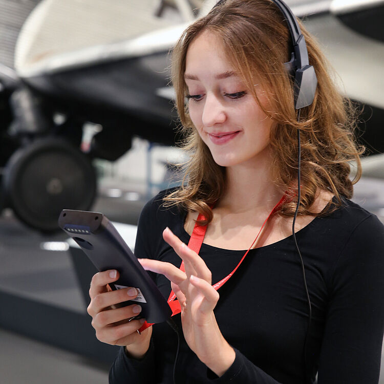 Eine Person mit braunen, schulterlangen Haaren trägt Kopfhörer und eine rote Besucherschleife, hält ein tragbares Audioguide-Gerät und tippt darauf, im Hintergrund Teile eines Flugzeugs in einem Museum.