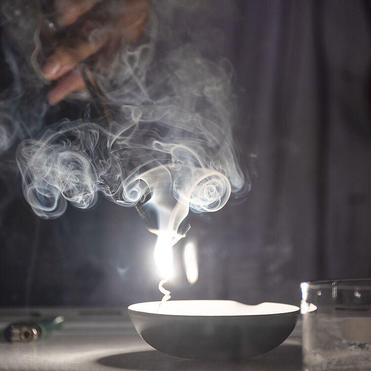 Eine Mitarbeiterin der Ausstellung Chemie zündet im Labor Salze an und eine weiße Flamme und Rauch sticht empor.