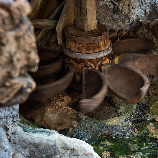 Nahaufnahme eines rotierenden Holzrad- oder Schaufelmechanismus in einer steinigen Wasserumgebung, verwitterte hölzerne Nabe mit Metallbändern und bewegten Schaufeln über grünlich bewachsenem Wasser und Kalkstein.
