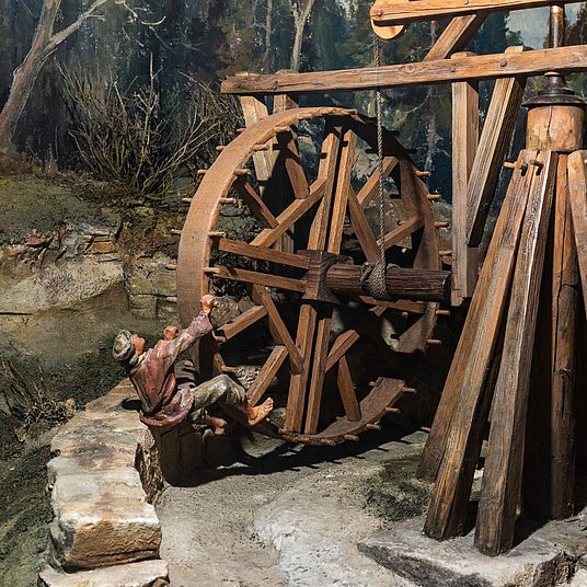 Nahaufnahme eines historischen hölzernen Wasserrads in einer Miniaturszene: großes, verwittertes Zahnrad mit Speichen, Seilbefestigung und Holzkonstruktion, daneben ein hölzerner Pfostenstapel, Steinmauer und eine kleine bemalte Figur eines Mannes, der am Rad hängt, vor einem bewaldeten Diorama-Hintergrund.