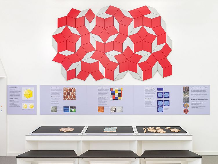 Drei Mitmach-Stationen zum Thema Parkettierungen. An der Wand befindet sich ein Ausschnitt einer Penrose-Parkettierung.