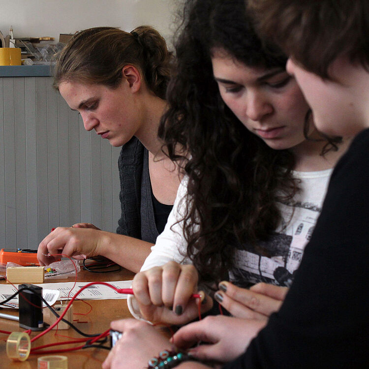 Drei Personen sitzen an einem Holztisch und arbeiten an Elektronik: sichtbare Drähte, Krokodilklemmen, ein Messgerät und Klebeband.